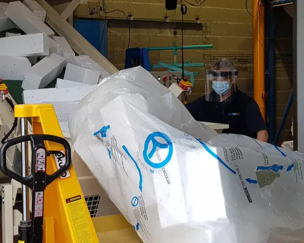 Pracownik w masce i przyłbicy przygotowuje styropian do recyklingu. Duży worek z logo Knauf Circular symbolizuje proces recyklingu styropianu.
