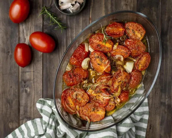 Pomodori gratinati al forno, dorati e succosi, con cipolle e rosmarino in una teglia di vetro su un tavolo di legno.