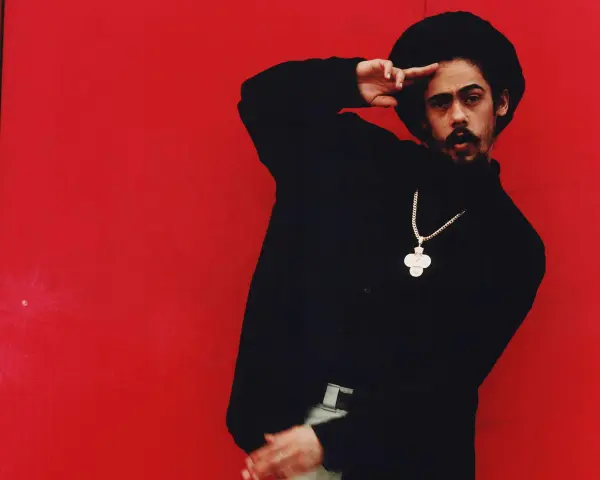 Mann mit Afro und Schnurrbart, schwarzer Rollkragenpullover, goldene Kette mit Kreuzanhänger, vor rotem Hintergrund. Er salutiert. "I shot the sheriff" könnte man sagen.