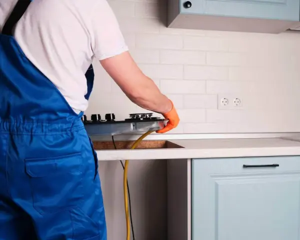 Instalator w niebieskim kombinezonie podłącza kuchenkę gazową w kuchni.