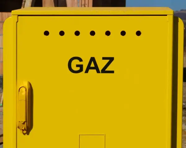 Żółta szafka gazowa z napisem "GAZ" i otworami wentylacyjnymi. Obok budowa domu, w tle krajobraz. Przyłącze gazowe krok po kroku.