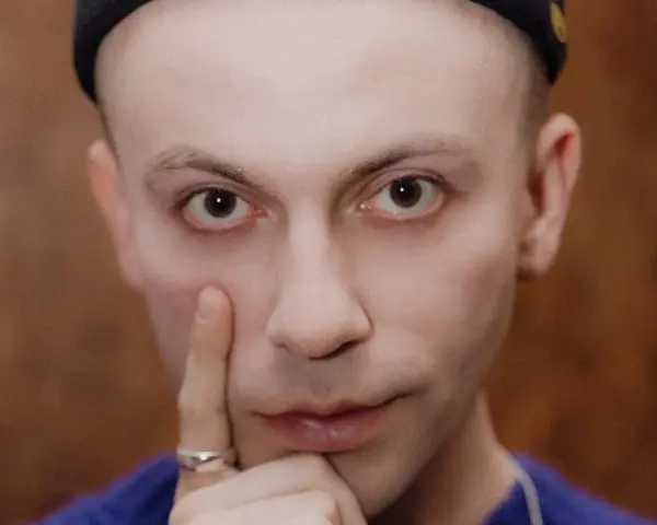 Zbliżenie na twarz Arka Kłusowskiego w czarnej czapce, z palcem przy policzku.