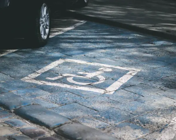 Niebieskie miejsce parkingowe dla niepełnosprawnych pod blokiem, z symbolem wózka inwalidzkiego. Obok widoczna część czarnego samochodu.