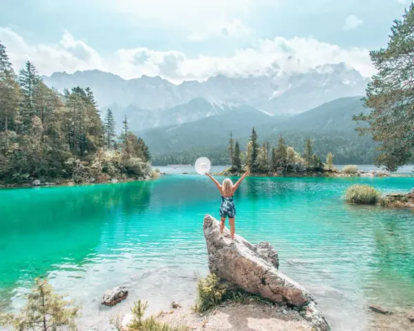 Frau mit ausgebreiteten Armen und Hut am türkisfarbenen Eibsee in Bayern, im Hintergrund Berge und Wald.