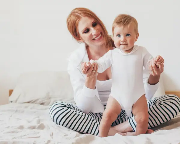 Uśmiechnięta mama pomaga swojemu 8 miesięcznemu dziecku w pierwszych krokach na łóżku.
