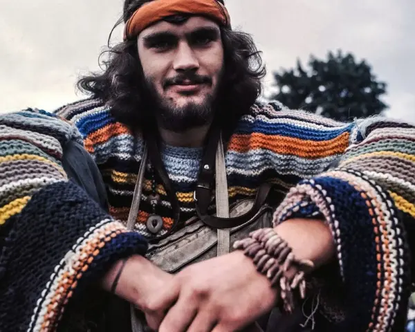 Młody mężczyzna w kolorowym swetrze w paski i opasce na głowie, uśmiecha się. Wygląda jak prawdziwy hippie.