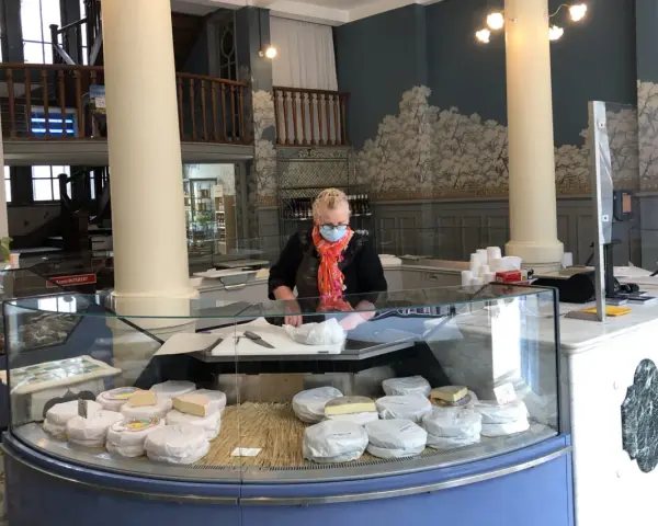 Une fromagère masquée prépare des fromages dans une vitrine réfrigérée.