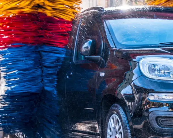 Czarna, błyszcząca karoseria samochodu w myjni automatycznej, otoczona kolorowymi szczotkami. Czy myjnia automatyczna niszczy lakier? Woda spływa po aucie.