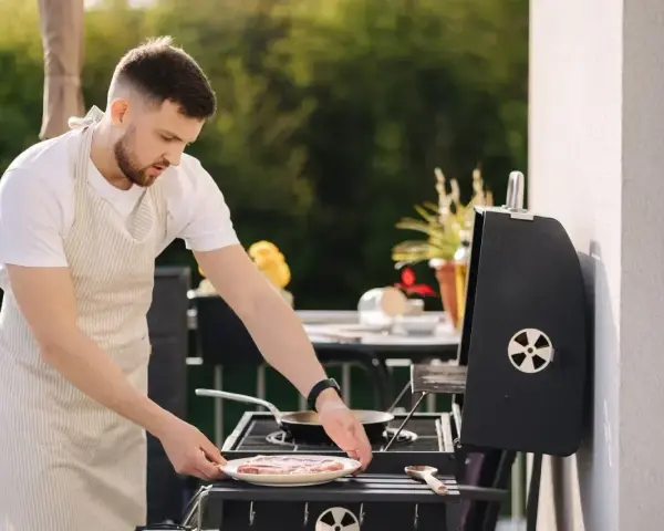 Mężczyzna w fartuchu przygotowuje mięso na grillu na balkonie. Czy można robić grilla na balkonie? Tak, jeśli jest bezpiecznie!