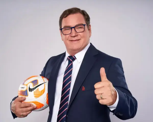 Uśmiechnięty trener reprezentacji Polski w garniturze trzyma piłkę i pokazuje kciuk w górę.