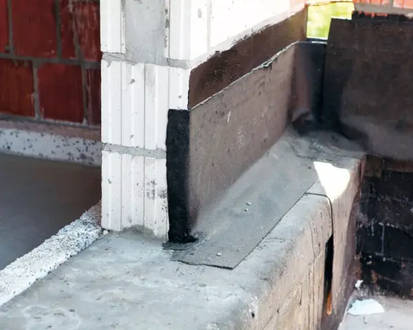 Fragment budowy z widoczną izolacją przeciwwilgociową, czyli papą na fundament. Widać beton, pustaki i czarną masę izolacyjną.