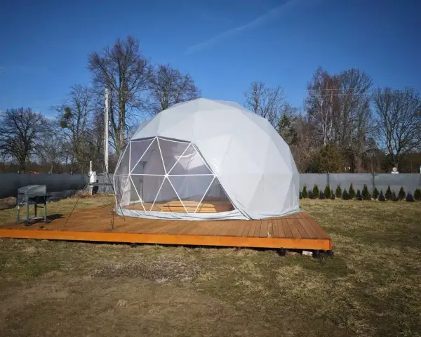 Nowoczesny glamping nad jeziorem. Biała kopuła na drewnianym tarasie, obok grill. Idealne miejsce na relaks.