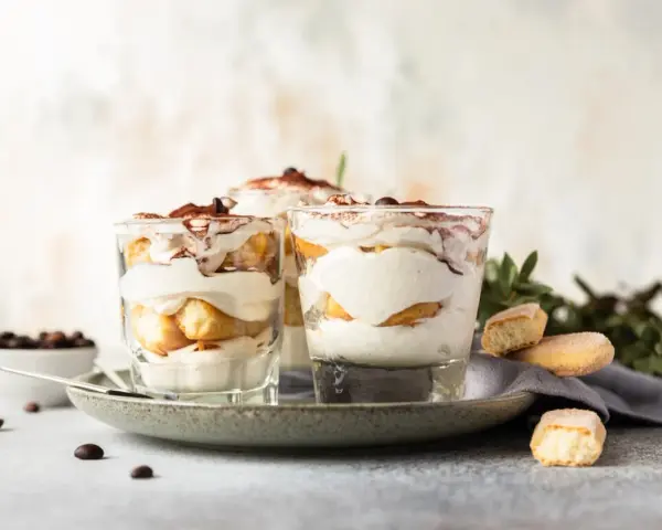Pyszne desery włoskie: tiramisu w szklankach, udekorowane kakao i kawą. Obok kawałki biszkoptów i ziarna kawy.