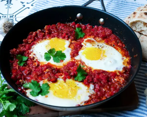 Herzhaftes Shakshuka-Rezept in einer Pfanne mit pochierten Eiern in würziger Tomatensoße, garniert mit Koriander. Dazu gibt es Fladenbrot.