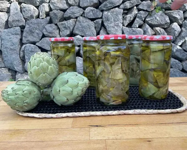 Alcachofas frescas y en conserva en frascos, sobre una mesa de madera y fondo de piedra.