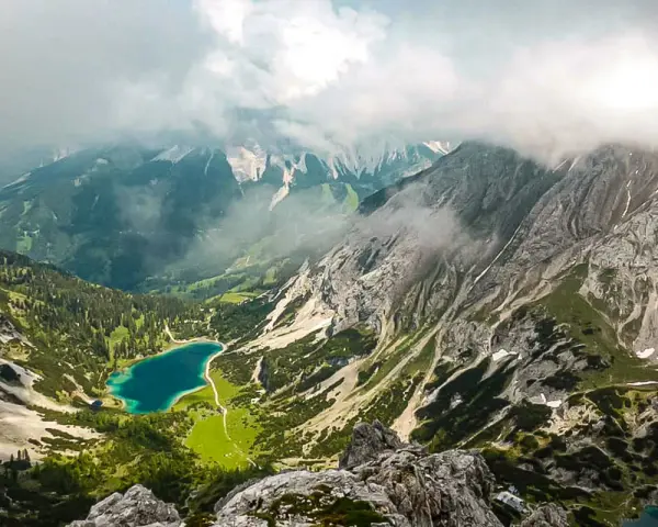 Blick vom Drachenkopf auf einen türkisfarbenen Bergsee, umgeben von grünen Hängen und felsigen Gipfeln, Wolken ziehen über die Landschaft.