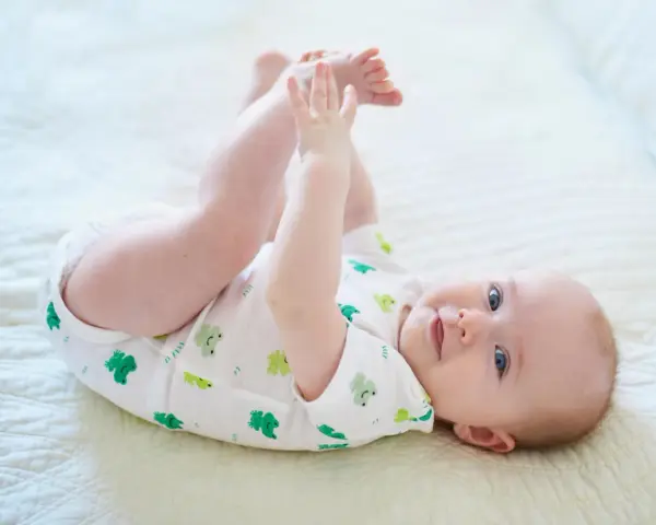 Ein Baby im 6. Lebensmonat liegt auf dem Rücken und spielt mit seinen Füßen. Es trägt einen weißen Body mit grünen Fröschen.