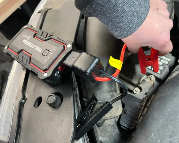 A hand connects red jumper cables from "The Athena" lithium jump starter to a car battery, demonstrating how lithium jump starters work by providing a temporary power boost.