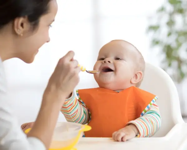 Mama karmi uśmiechniętego niemowlaka łyżeczką. To idealny przykład, co na śniadanie dla niemowlaka – zdrowo i radośnie!