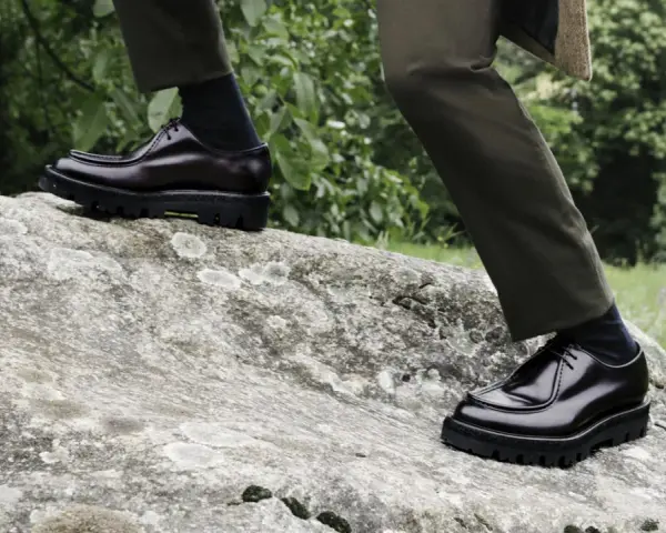 Scarpe oxford derby lucide su una roccia, con pantaloni verdi e sfondo verdeggiante.
