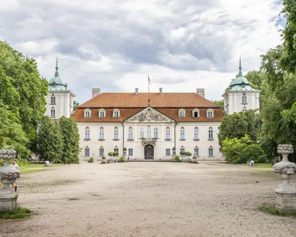 Pałac w Nieborowie, biała rezydencja z czerwonym dachem, otoczona zielenią. Dwie wieżyczki z zielonymi kopułami po bokach.