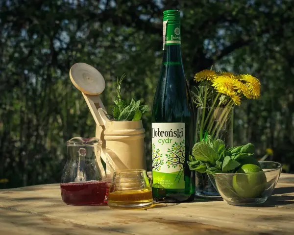 Butelka Dobroński cydr, dzbanuszek z czerwonym napojem, szklanka z bursztynowym płynem, limonki, mięta i żółte kwiaty na drewnianym stole.