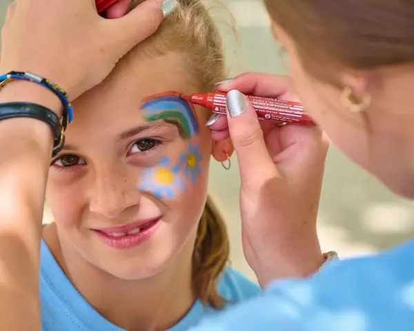 Une petite fille souriante se fait maquiller sur la joue un arc-en-ciel et des fleurs, au stand maquillage d'une kermesse.