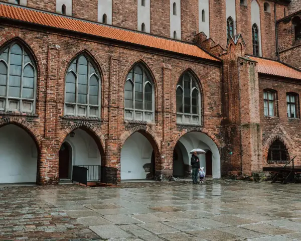 Zamek w Kwidzynie, z ceglanymi arkadami i gotyckimi oknami, skrywa tajemnice. Dwoje ludzi z parasolem spaceruje po mokrym dziedzińcu.