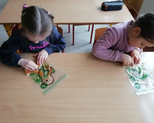 Dwie dziewczynki bawią się materiałami sensorycznymi w woreczkach na drewnianym stole.