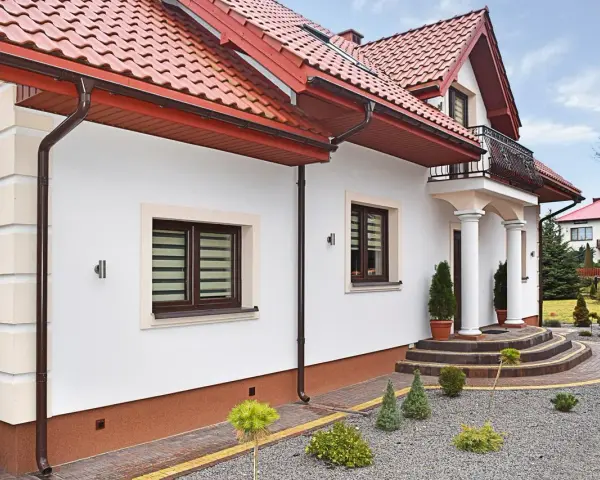 Elegancki dom z białym tynkiem i brązowymi detalami. Cokół wykończony ozdobnym tynkiem mozaikowym dodaje mu charakteru.