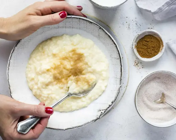 Cremiger Milchreis mit Vanillepudding, bestreut mit Zimt. Eine Hand mit roten Nägeln hält einen Löffel bereit.