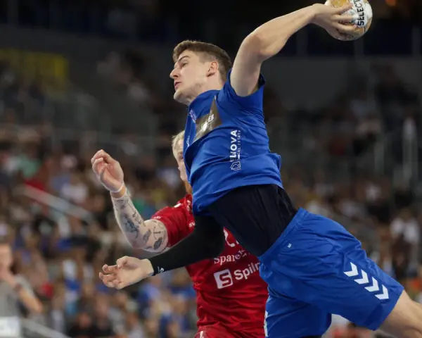 Magnus Landin Jacobsen w skoku do rzutu piłką ręczną, w niebieskim stroju, z widownią w tle.