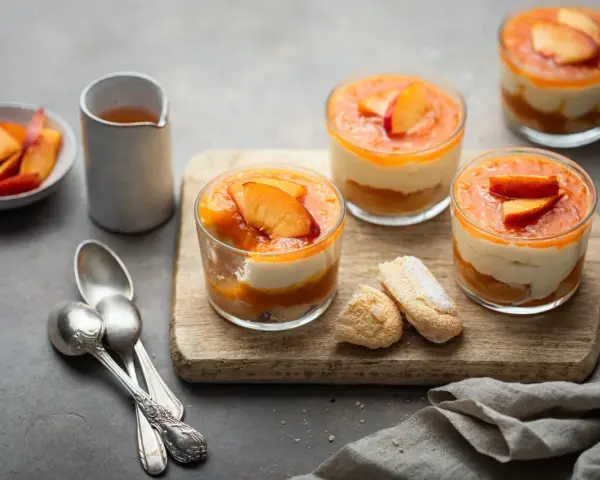 Delizioso tiramisù alle pesche in coppette, con pesche fresche e savoiardi su tagliere di legno.