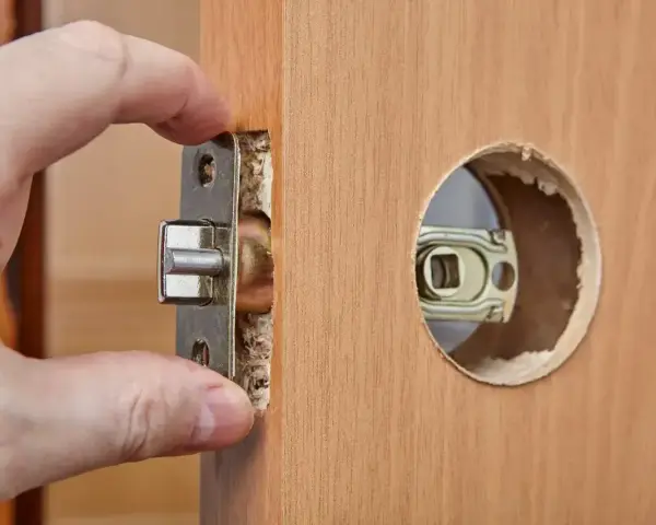 Dłoń montuje mechanizm zamka w drewnianych drzwiach. Dowiedz się, ile kosztuje wymiana zamka w drzwiach.