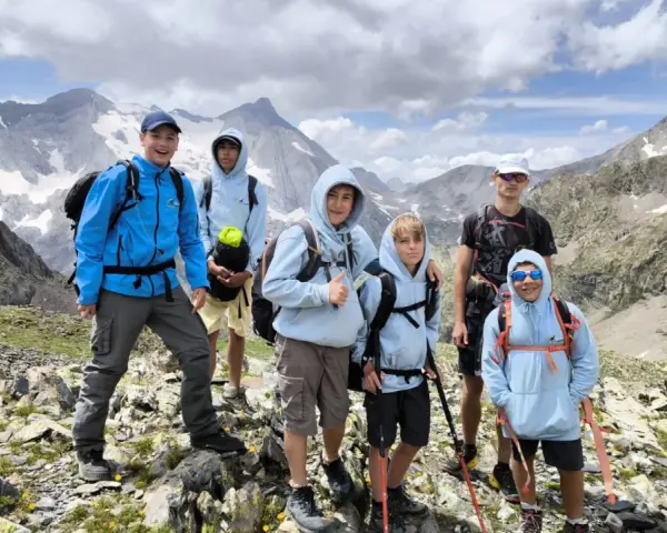 Groupe d'enfants et d'un adulte en randonnée en montagne, souriants, avec des sacs à dos et des bâtons de marche.