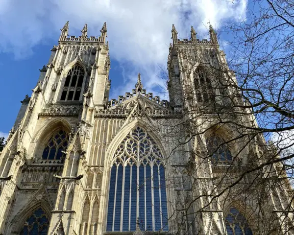 The majestic York Minster, a Gothic cathedral, stands tall against a bright blue sky with scattered clouds. Bare tree branches frame the intricate stonework and towering spires.