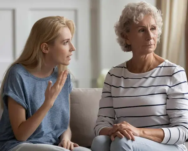 Junge Frau spricht vorwurfsvoll zu ihrer Mutter, die abgewandt sitzt. Die Mutter beleidigt die erwachsene Tochter.