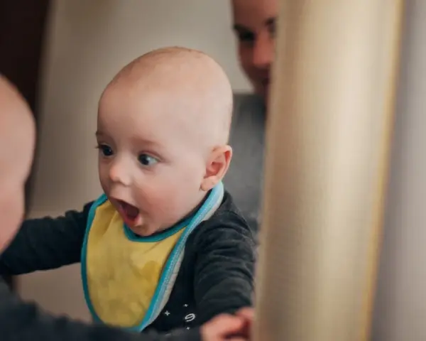 Ein Baby mit gelbem Lätzchen schaut überrascht in den Spiegel. Es ist fasziniert von seinem Spiegelbild, ein Moment, wann sagen Kinder "Ich".