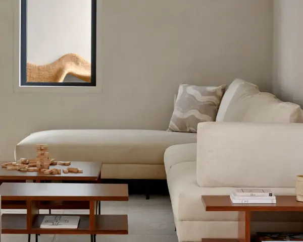 Sofá modular beige, mesa de centro de madera con juego de Jenga, planta en maceta y ventana con detalle decorativo. Ambiente de estilo cozy.