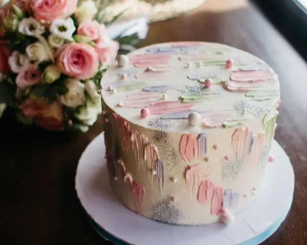 Oto tort na 20 osób, ważący około 2 kg, ozdobiony pastelowymi pociągnięciami pędzla i perełkami. Obok bukiet różowych kwiatów.