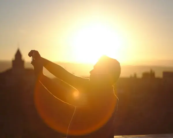 Silhouette d'une personne prenant une photo contre jour avec son téléphone, le soleil éclatant à l'horizon derrière des bâtiments.