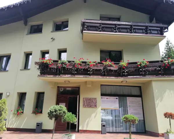 Jasnozielony hotel na błoniach z balkonem pełnym czerwonych kwiatów.