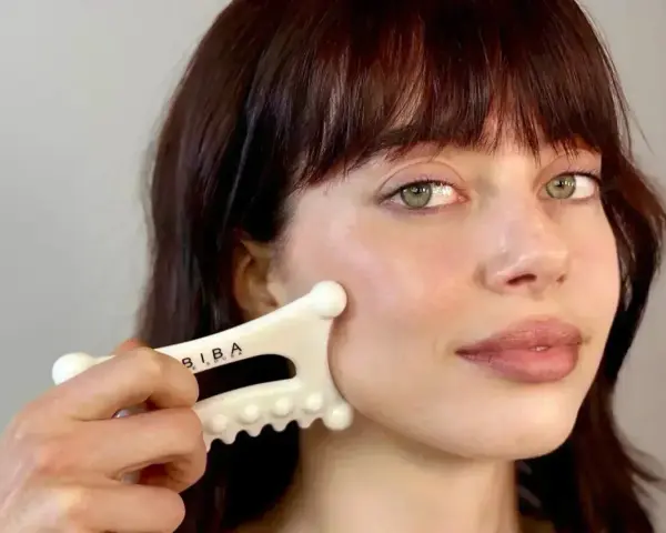 A woman with bangs and green eyes performs a facial fascia massage using a white gua sha tool.