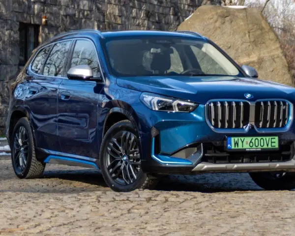 Niebieskie BMW iX1 stoi na kamienistej nawierzchni przed kamienną ścianą.