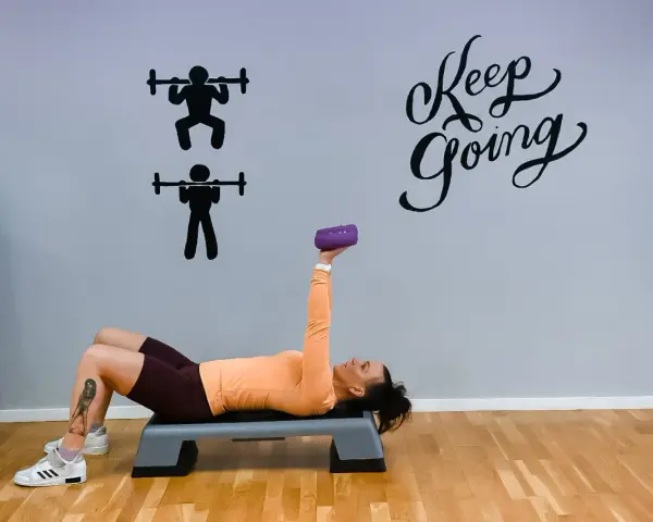Kvinna tränar med en lila hantel på en stepbräda. Väggen har texten "Keep Going" och silhuetter av styrkelyft.