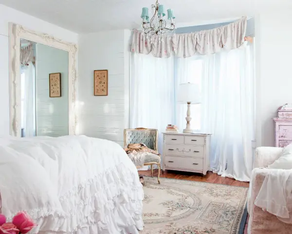 Dormitorio con cama blanca y volantes, espejo grande, cómoda antigua y sillón. Estilo shabby chic.