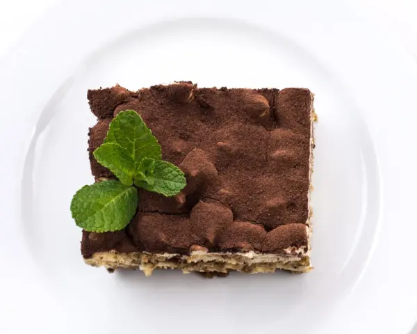 Pyszne tiramisu bez alkoholu, posypane kakao i udekorowane listkami mięty, na białym talerzu.