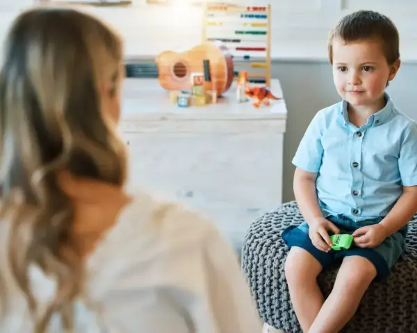 Chłopiec siedzi na pufie naprzeciwko psychoterapeutki, w tle zabawki.