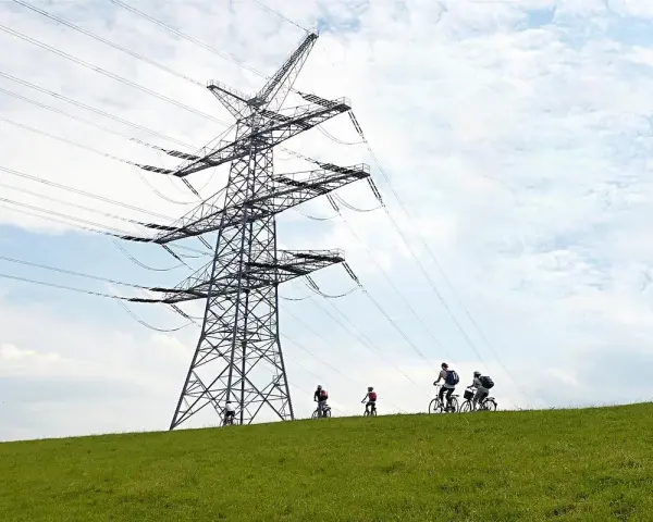 Grupa rowerzystów przemierza zielone wzgórze pod potężną konstrukcją linii 110 kV.