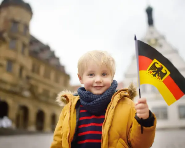 Uśmiechnięty chłopiec z blond włosami trzyma flagę Niemiec. W tle widać zabytkowe budynki, przypominające te, które można spotkać w niemieckich miasteczkach, gdzie znajdują się szkoły.
